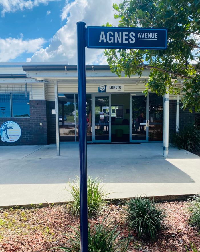Sign reading “Agnes Avenue” in front of a school entrance with glass doors and a Loreto sign above.