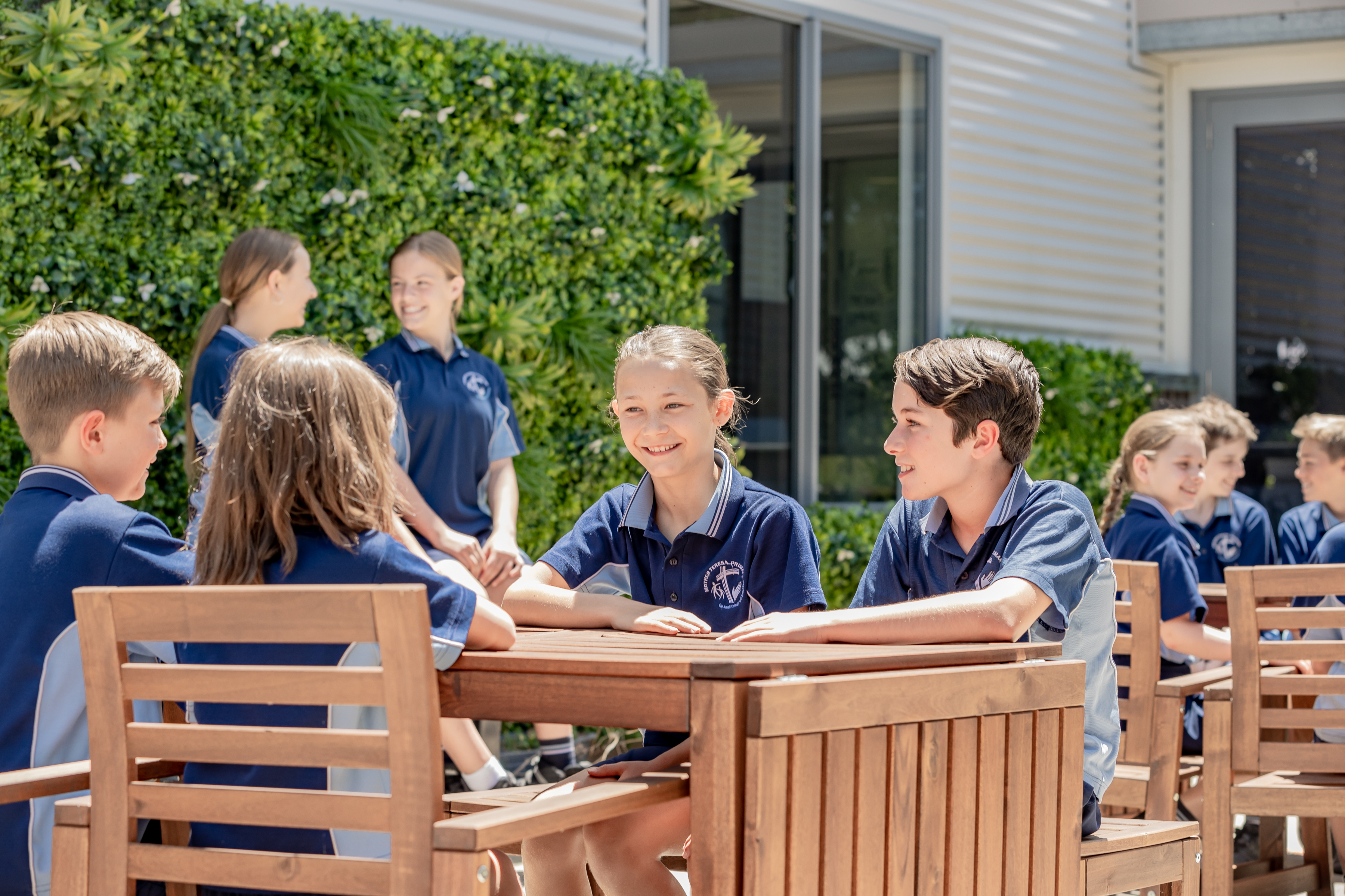 Students in school uniforms sitting and talking together at outdoor tables on a school campus.