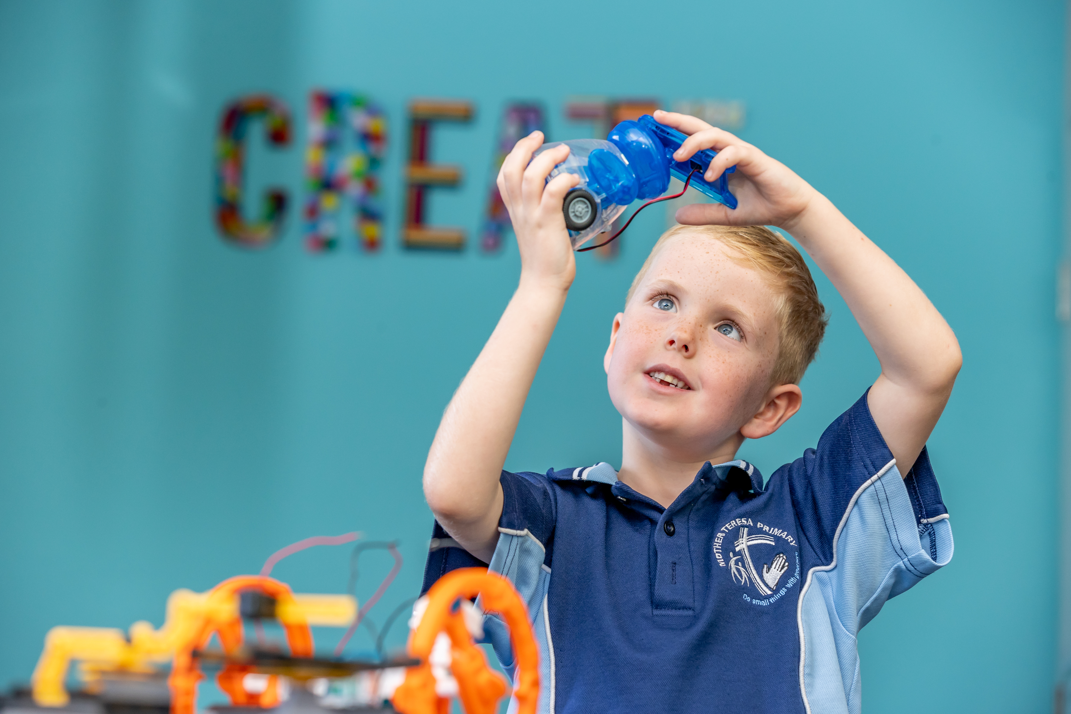 Child holding up a small wheeled device made with a plastic bottle during a STEM activity, with a “CREATE” sign on the wall behind.