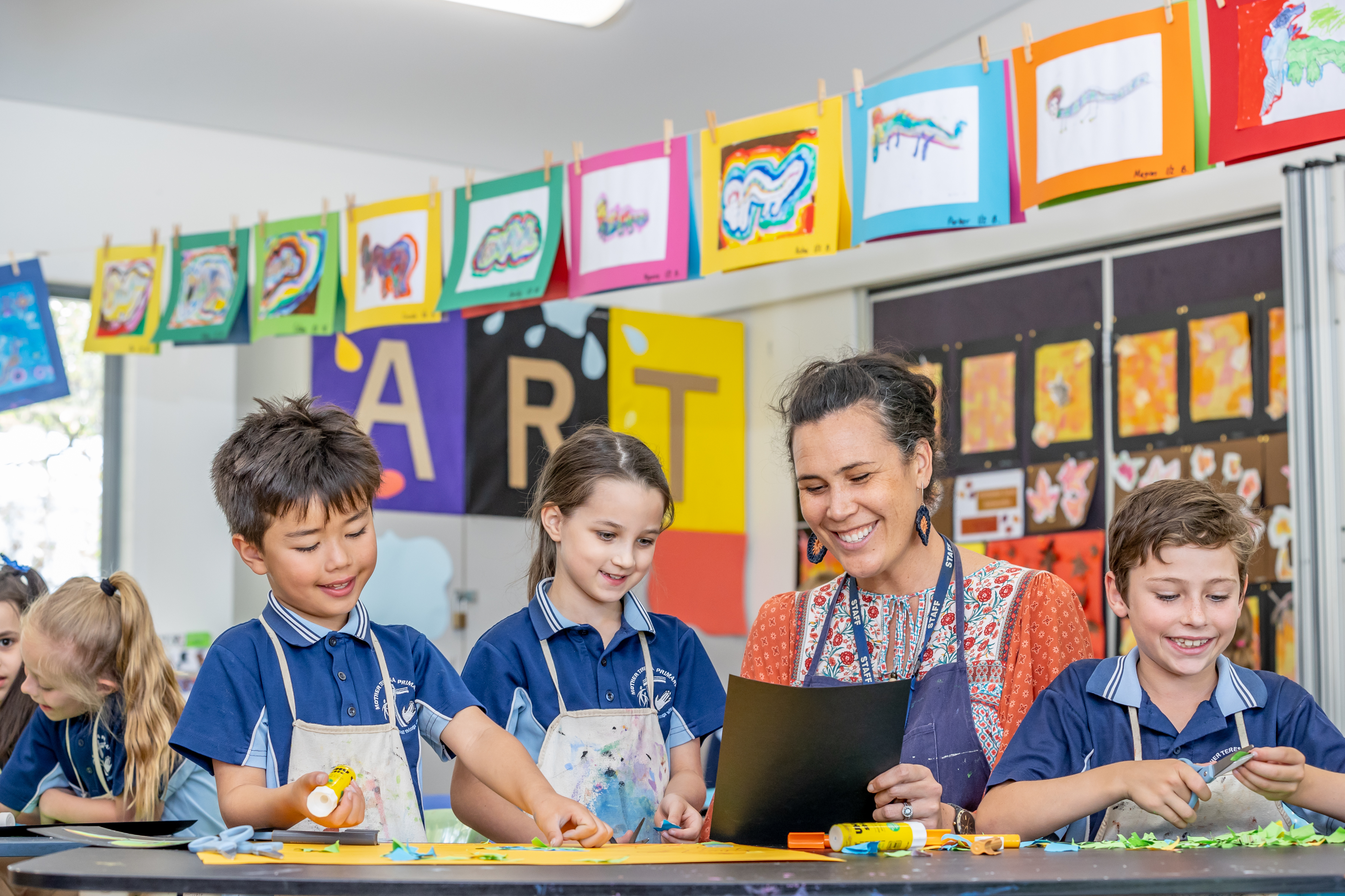Students and a teacher creating artwork with paper and glue in a classroom, with colourful art displays behind them.