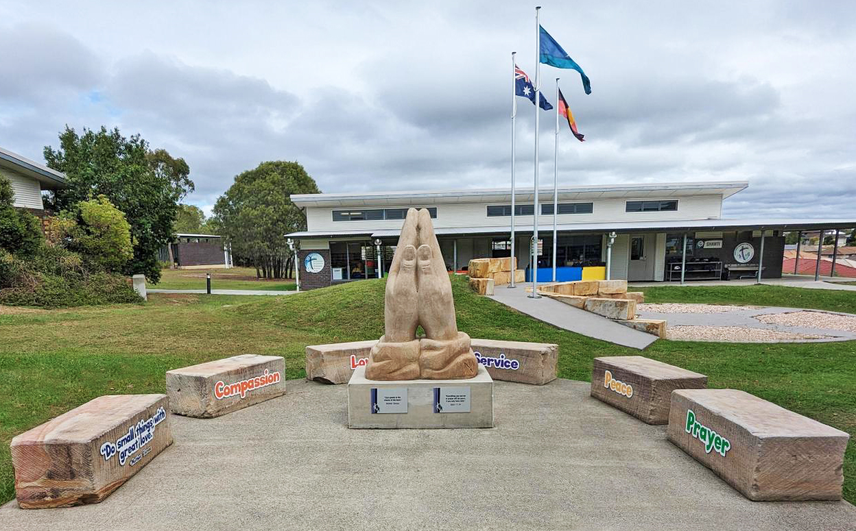 School courtyard with a central sculpture of praying hands, surrounded by engraved stone blocks and flagpoles in front of buildings.